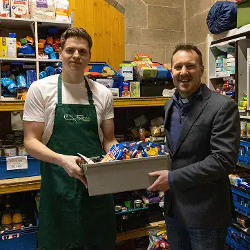 Two men in a food bank, one in a green apron and the other in a suit, exchanging a box of food.