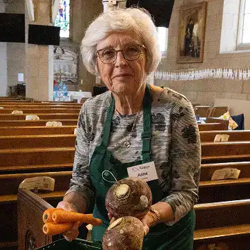 An elderly woman wearing glasses and a name badge, holding carrots and root vegetables inside a chur.