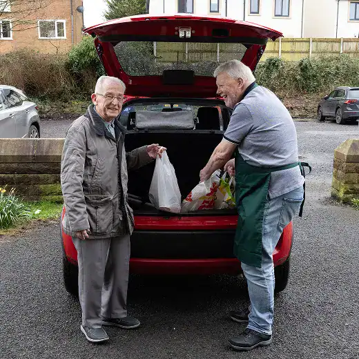 Volunteer handing food bags to a recipient from the open trunk of a red car.